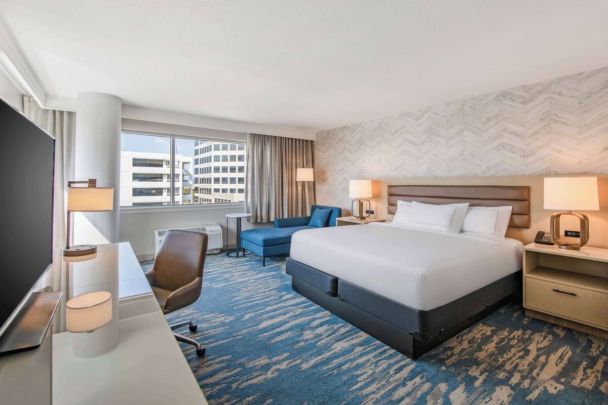 A hotel room with a large bed, a desk with a chair, a TV, and a view of the city through the window.