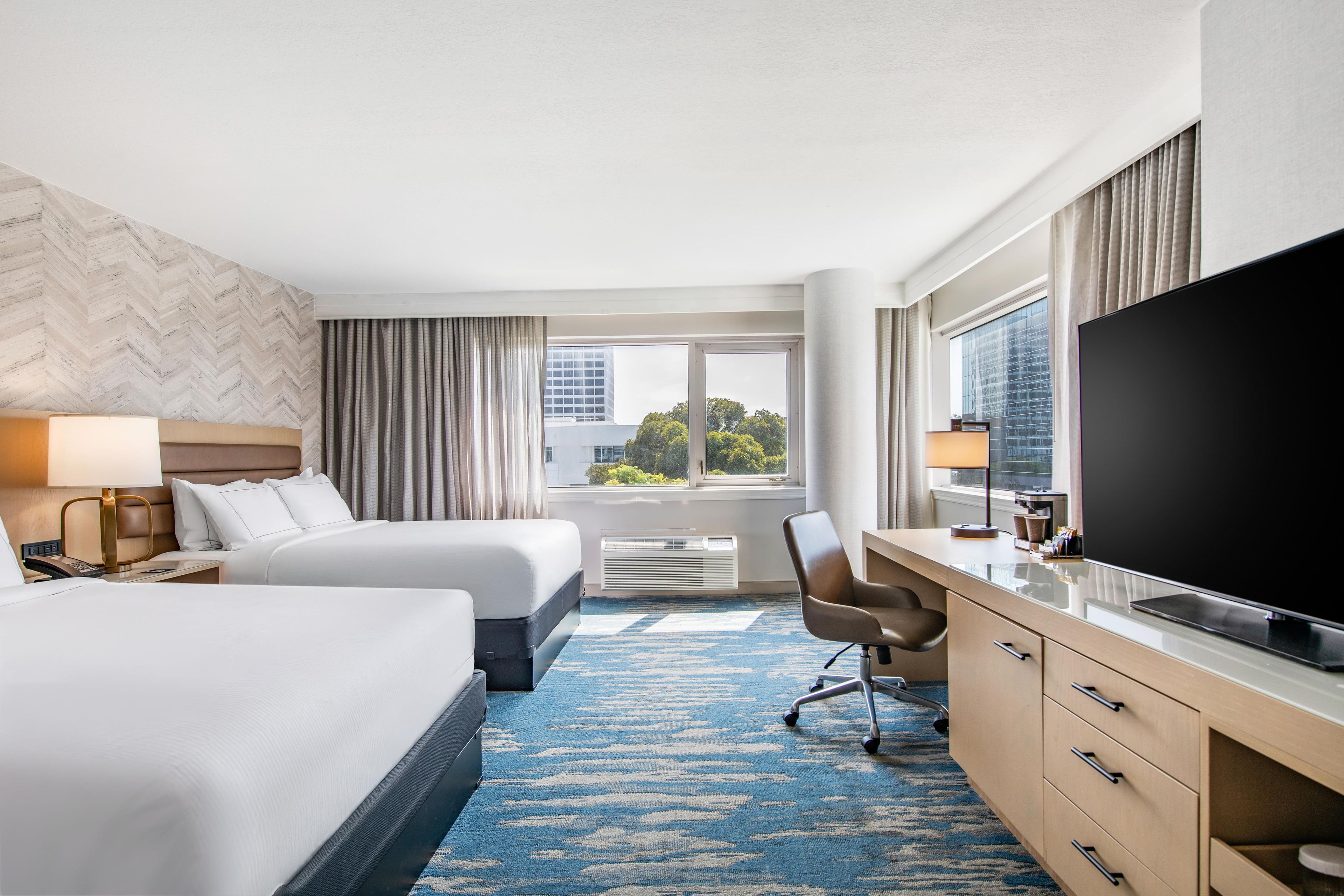 Ein Hotelzimmer mit zwei Betten, einem Schreibtisch, einem Flachbildfernseher, einem Sessel und einem großen Fenster mit Blick auf die Stadt.
