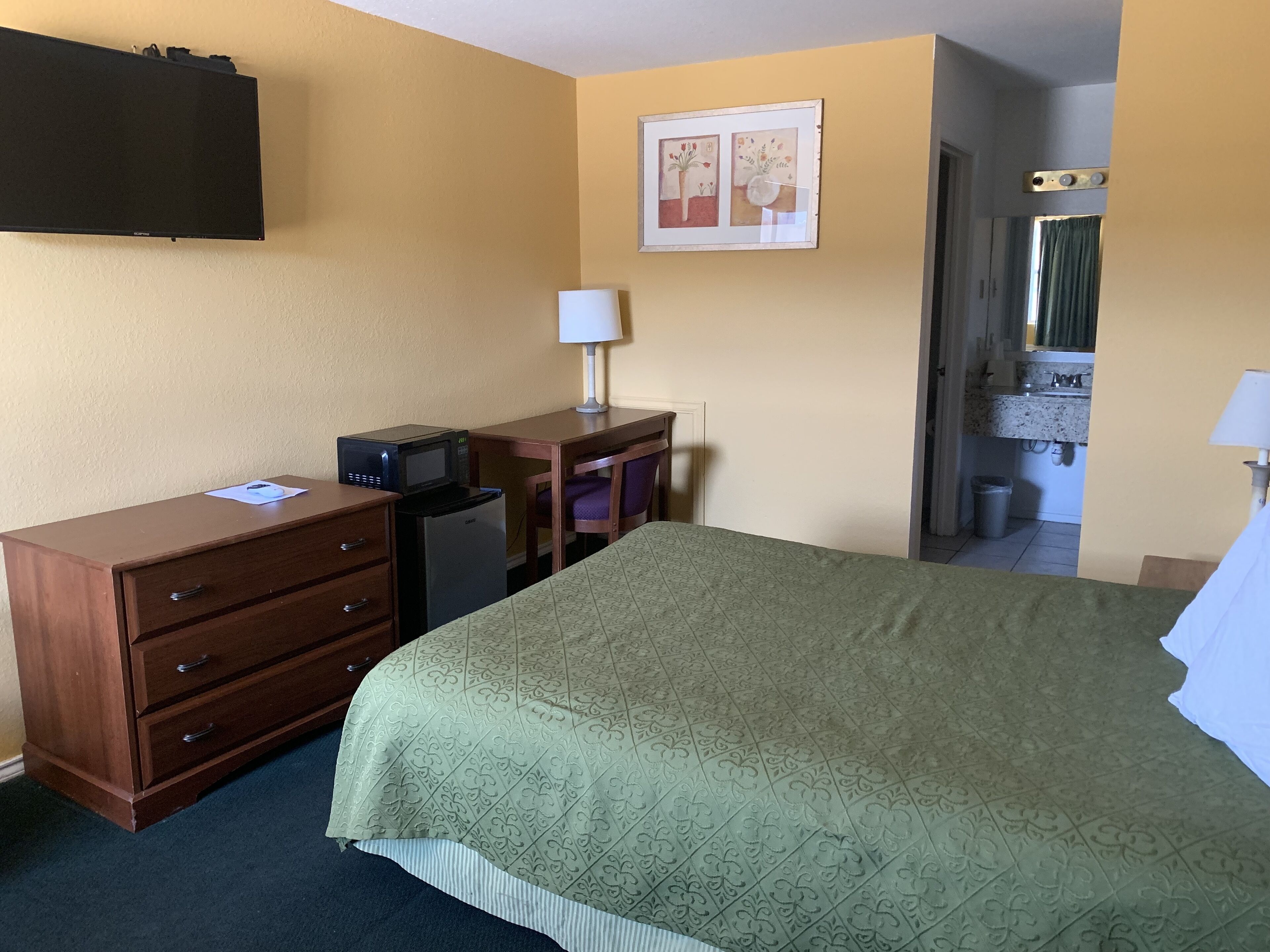 Une chambre d’hôtel équipée d’un lit, d’un bureau avec un ordinateur, d’un four à micro-ondes, d’un réfrigérateur, d’une télévision et d’une salle de bain visible à travers une porte ouverte.