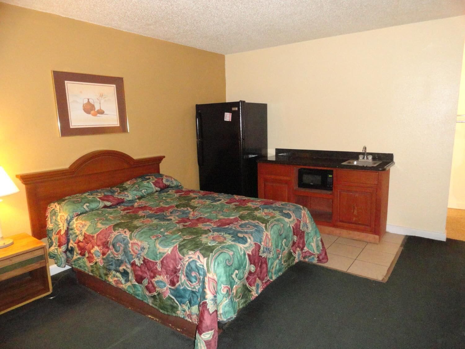 A hotel room with a bed, a small wooden nightstand, a refrigerator, a microwave, and a sink in the kitchenette.