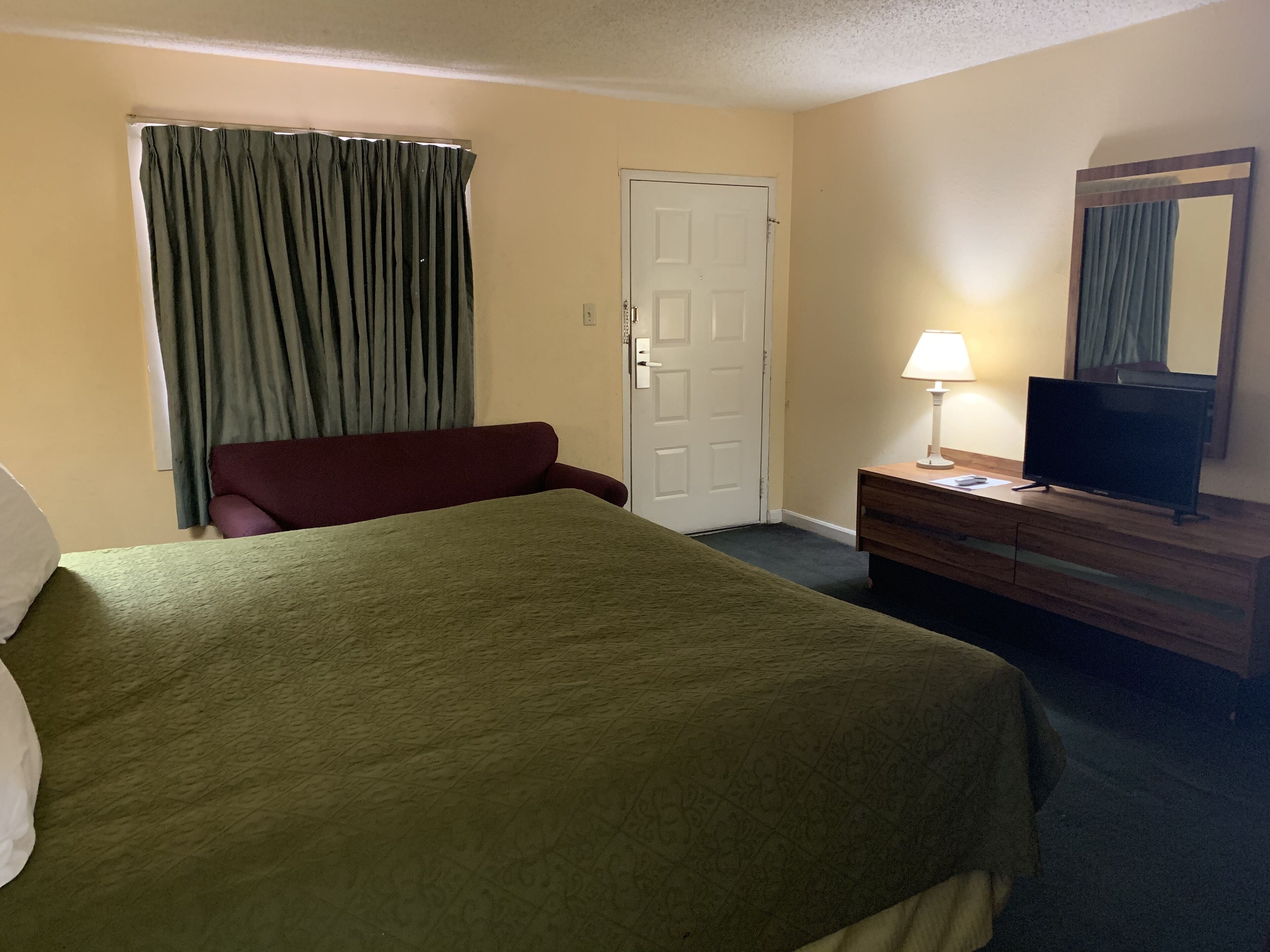Una habitación de hotel con una cama, un escritorio con lámpara, televisión y una puerta.