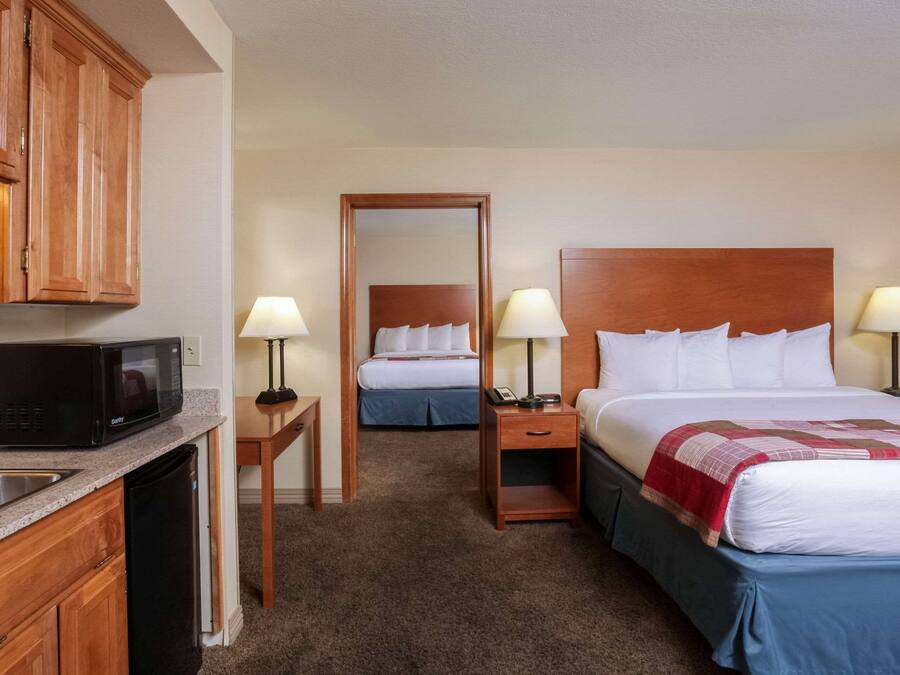 Una habitación de hotel con kitchenette, una cama con cabecera de madera, dos mesitas de noche con lámparas y un espejo de cuerpo entero.