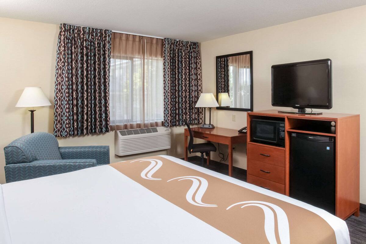 A hotel room with a bed, desk, TV, and kitchenette.