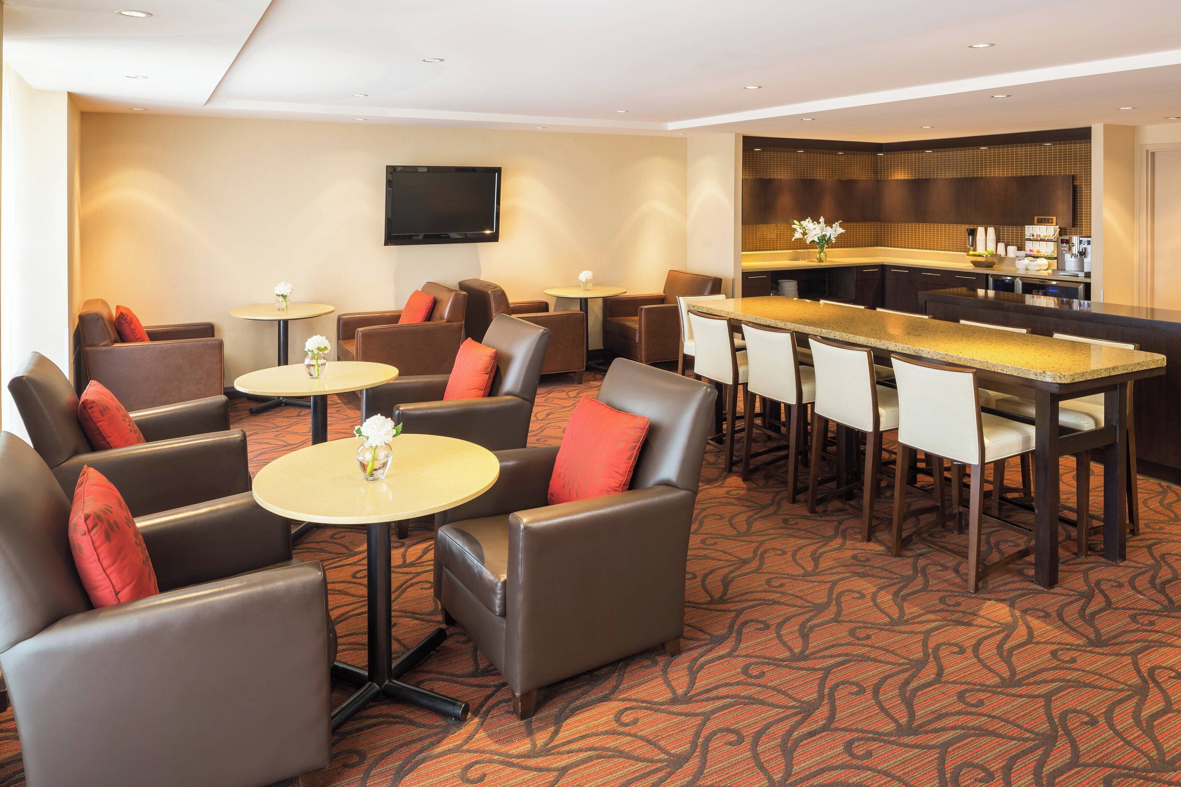 A hotel lobby with leather armchairs, round tables, a bar area, and a flat-screen TV on the wall.