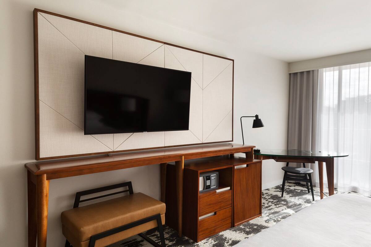 Habitación de hotel con un televisor de pantalla plana grande montado en una pared de paneles de madera, un escritorio de madera con una silla, un pequeño banco y una mesita de noche con una lámpara.