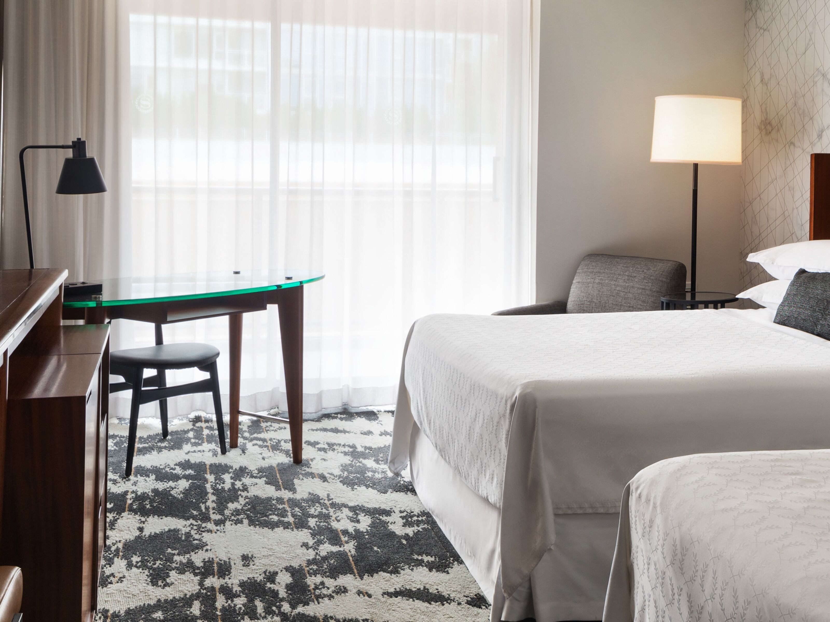 Una habitación de hotel con cama, escritorio, silla, lámpara y ventana con cortinas translúcidas.