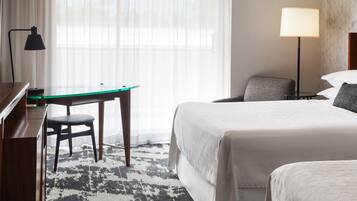 Una habitación de hotel con cama, escritorio, silla, lámpara y ventana con cortinas translúcidas.