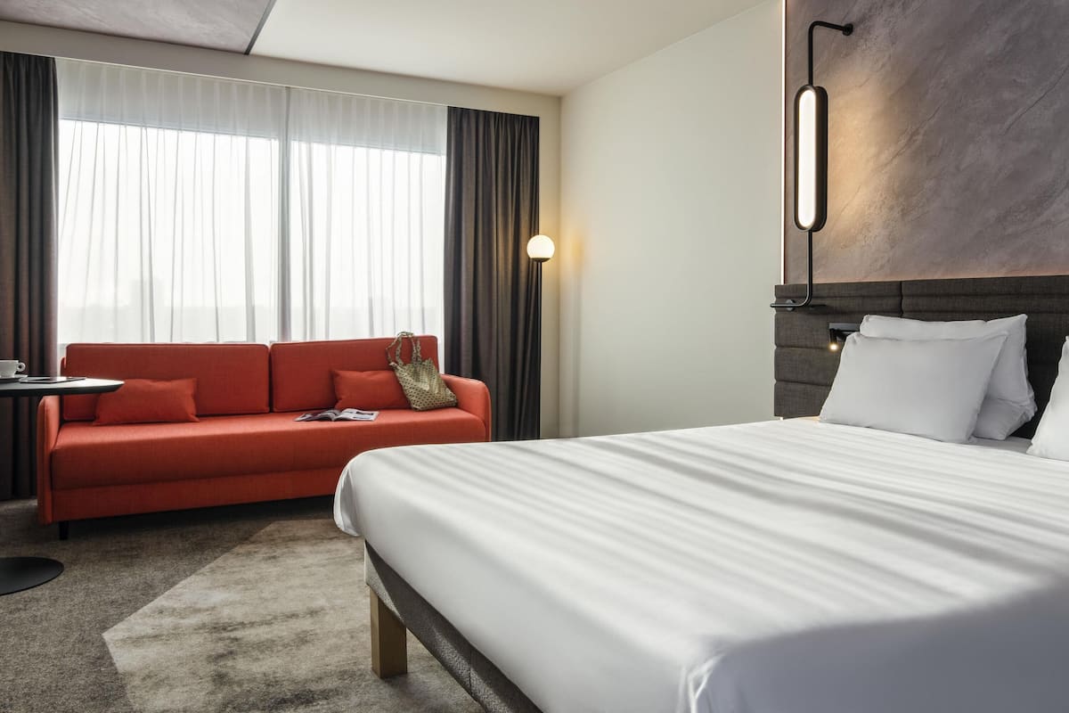 A hotel room with a bed, a red sofa, a small table with a cup, and a lamp.
