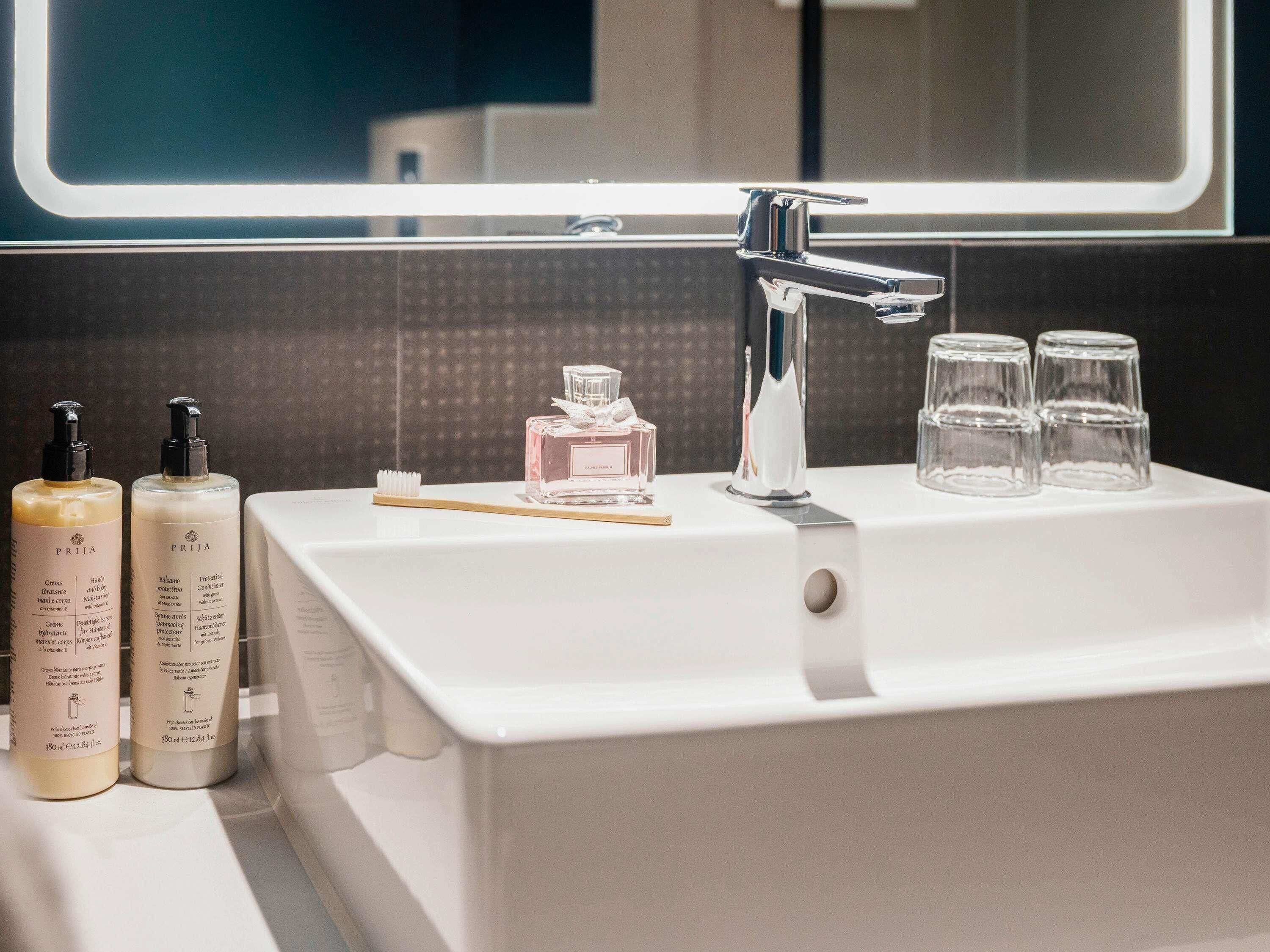A bathroom sink with a mirror, two bottles of shampoo, a perfume bottle, and three glasses.