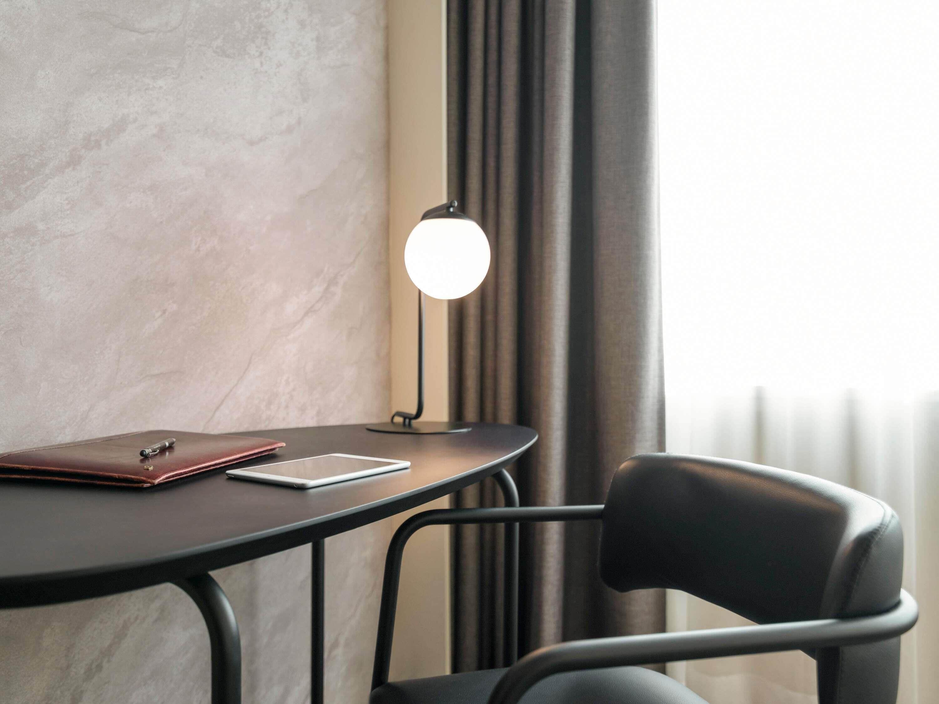 Ein Tisch mit einem Laptop, einem Notizbuch und einem Stift, eine Lampe und ein Stuhl in einem Raum mit strukturierter Wand und Vorhängen.