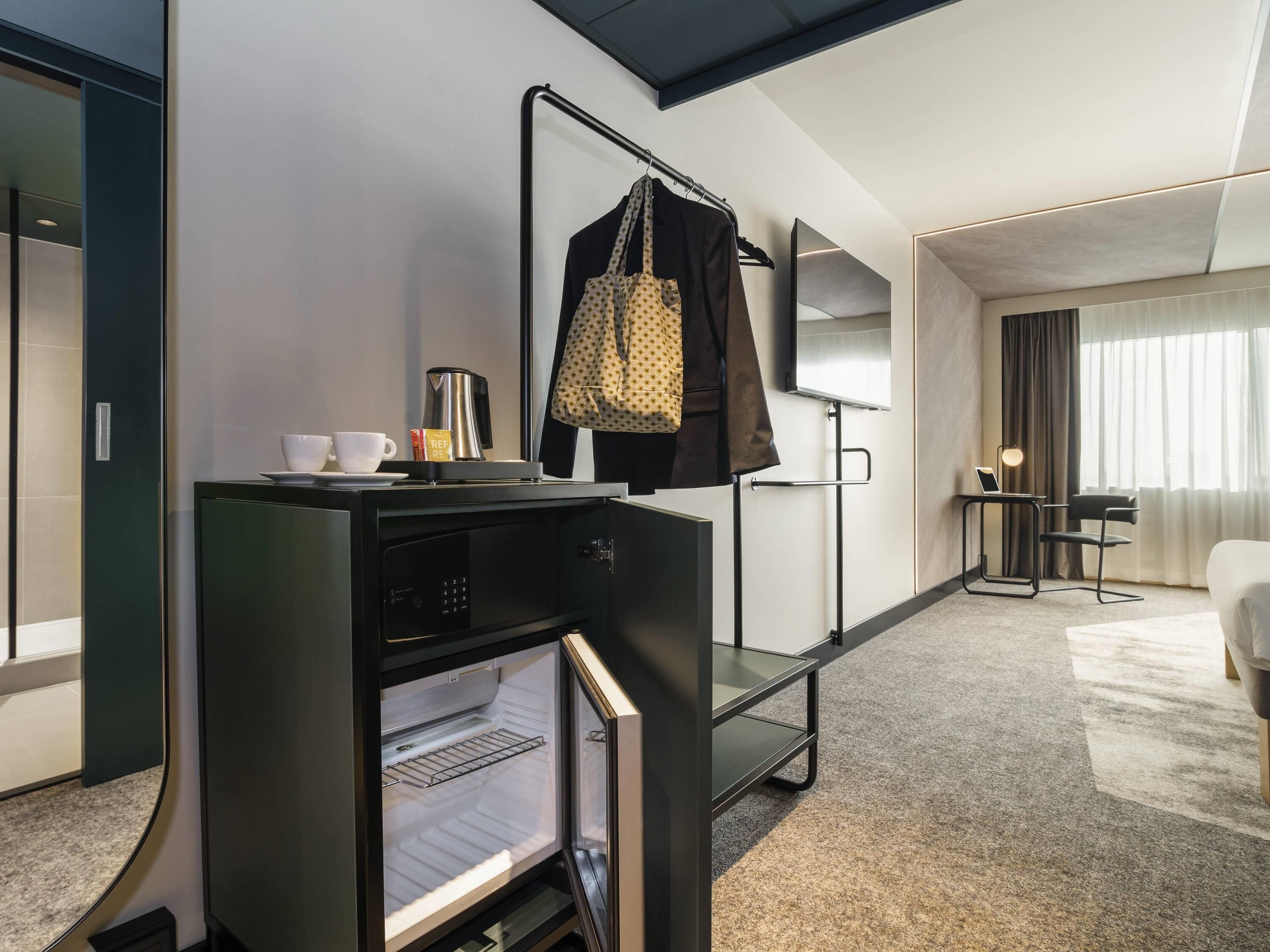 Habitación de hotel con minibar, cafetera, perchero, escritorio con computadora portátil y cama.