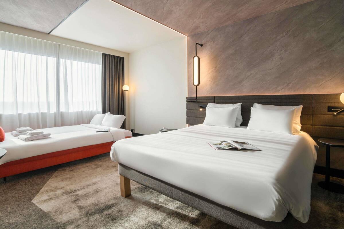 Une chambre d’hôtel avec un grand lit, un lit plus petit, une table de chevet, une lampe et une fenêtre avec des rideaux.