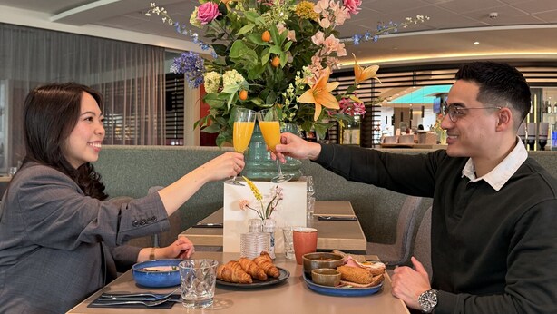 A dining table with a floral centerpiece, two people toasting, and breakfast items like croissants and orange juice.