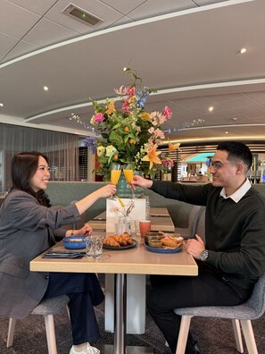 A dining table with a floral centerpiece, two people toasting, and breakfast items like croissants and orange juice. - Novotel Rotterdam Brainpark (Rotterdam)