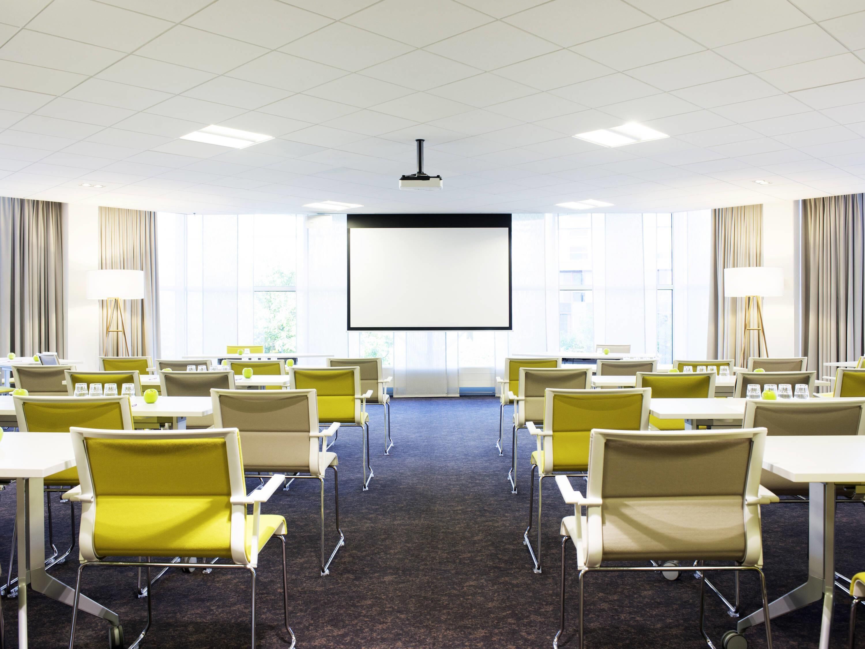 Une salle de conférence avec des rangées de chaises jaunes, un grand écran à l’avant et de grandes fenêtres équipées de rideaux.