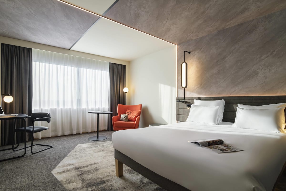 A modern hotel room with a large bed, a desk, a chair, and a lamp.