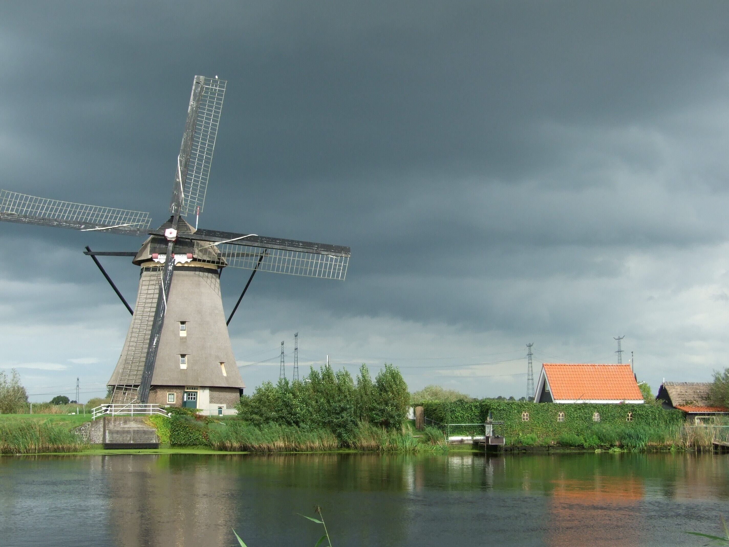 Un moulin à vent traditionnel, situé près d’une rivière paisible, avec une petite maison à proximité et un ciel nuageux au-dessus.