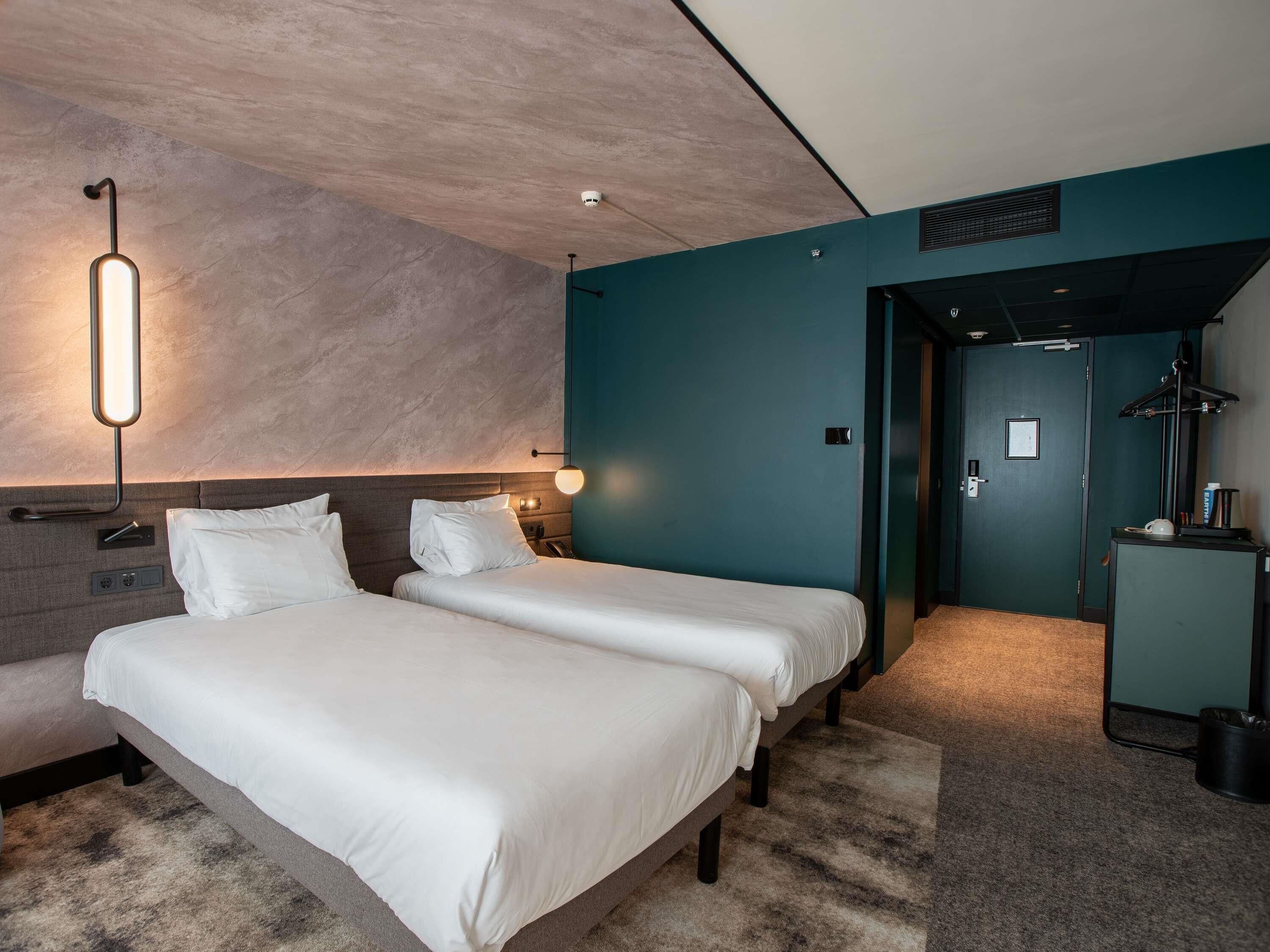 A hotel room with two beds, a desk, a chair, a lamp, and a door leading to another room.