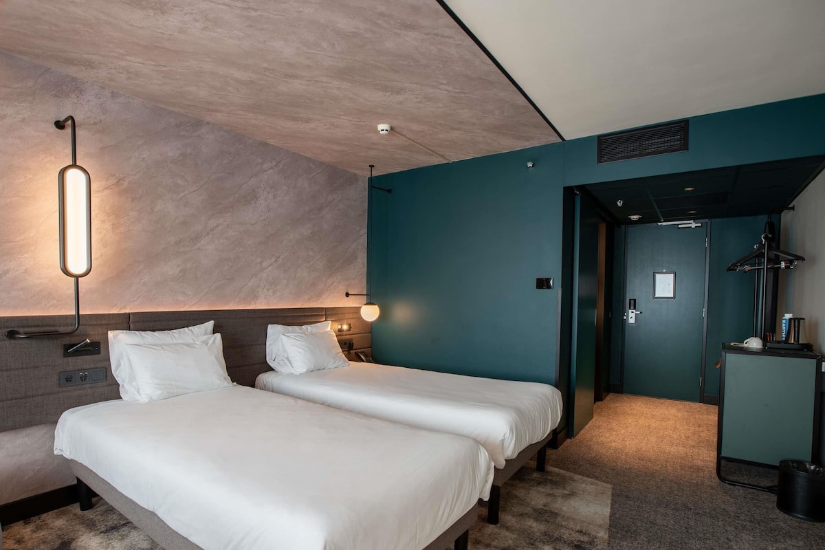 Ein Hotelzimmer mit zwei Betten, einem Schreibtisch, einem Stuhl, einer Lampe und einer Tür zu einem anderen Raum.