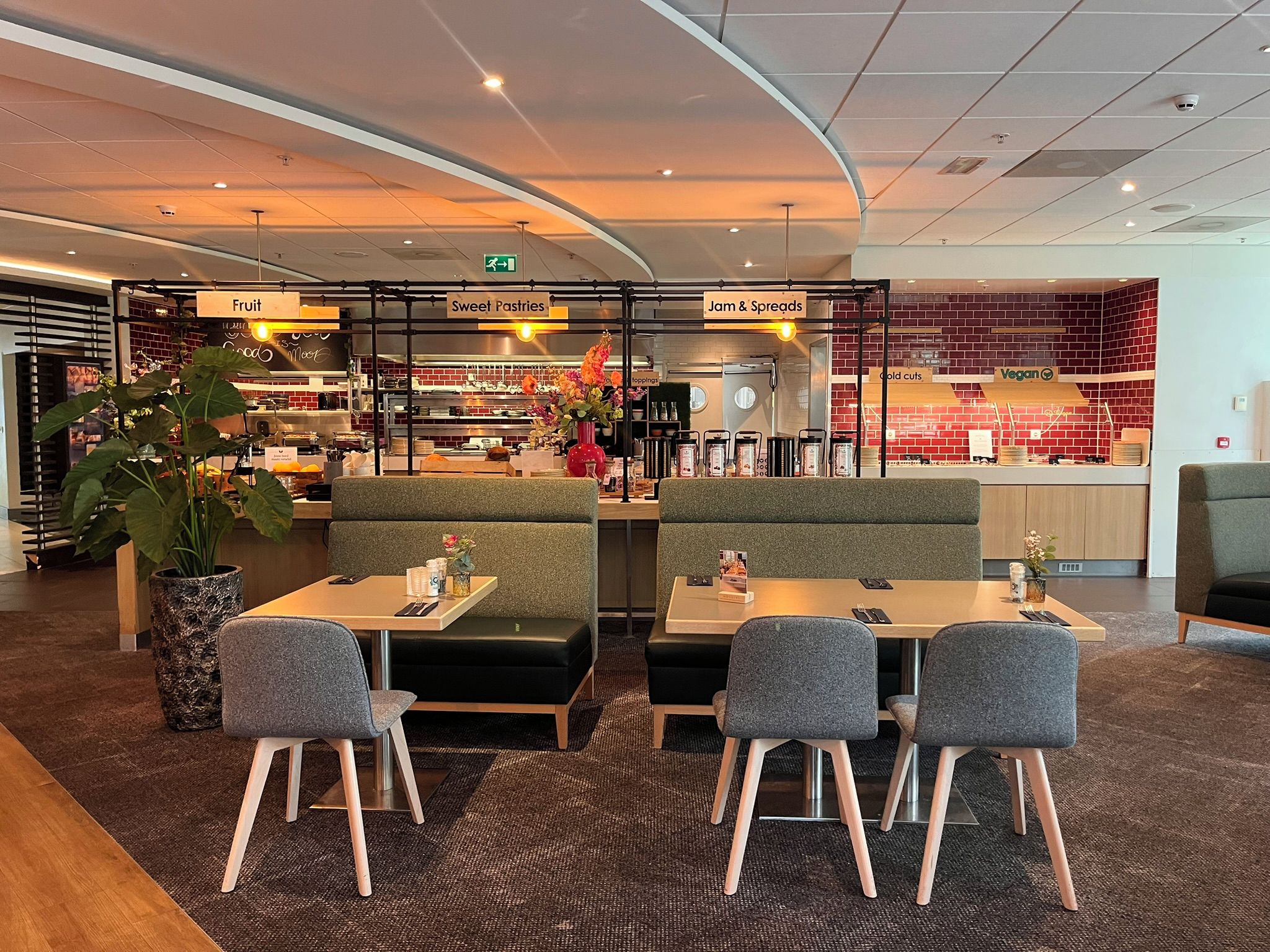 Un restaurant avec des banquettes et des tables, un mur en briques et des panneaux indiquant « Fruits », « Pâtisseries » et « Confitures et tartinades ».