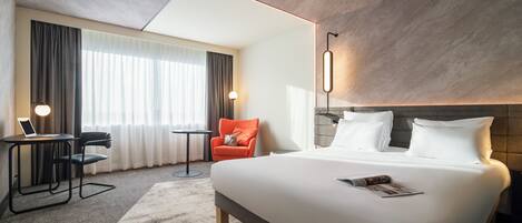 A modern hotel room with a large bed, a desk with a laptop, a chair, and a fireplace.