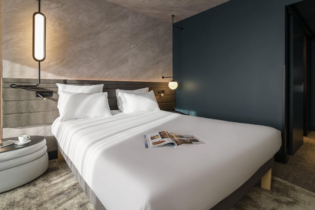 A modern hotel room with a large bed, a bedside table with a cup, and a magazine on the bed.