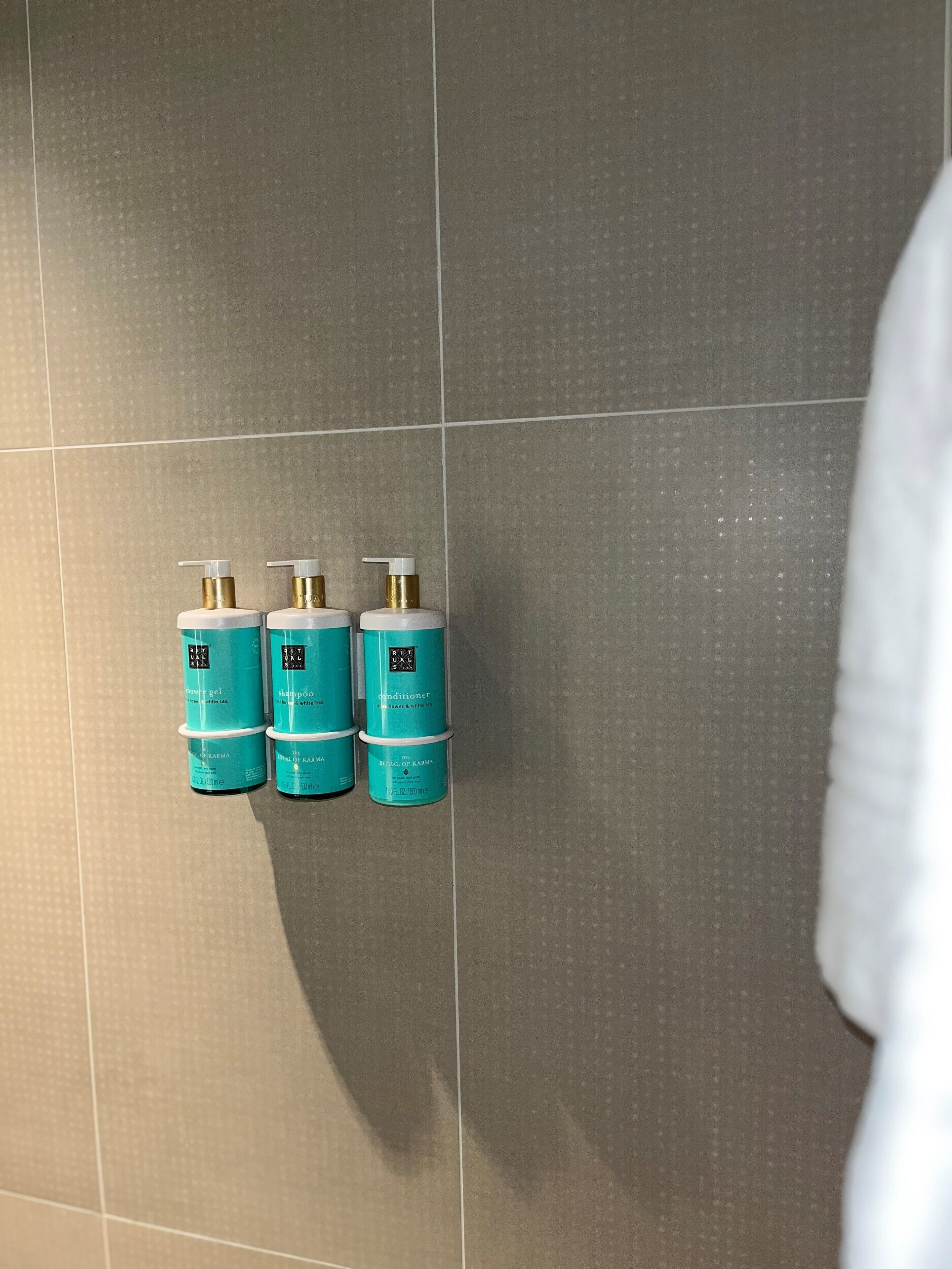 Três frascos azul-esverdeados de produtos Ritual of Karma sobre uma parede de azulejos.