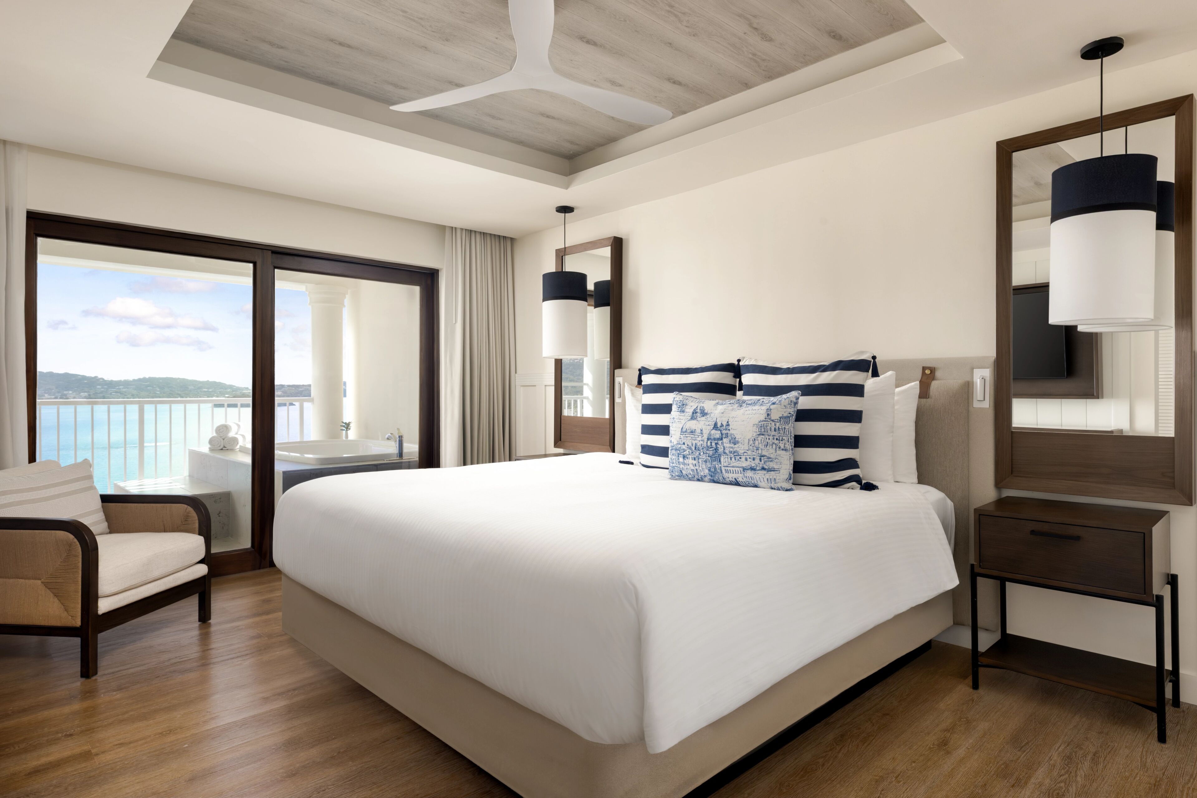 A hotel room with a large bed, a chair, a nightstand, a mirror, and a view of the ocean.