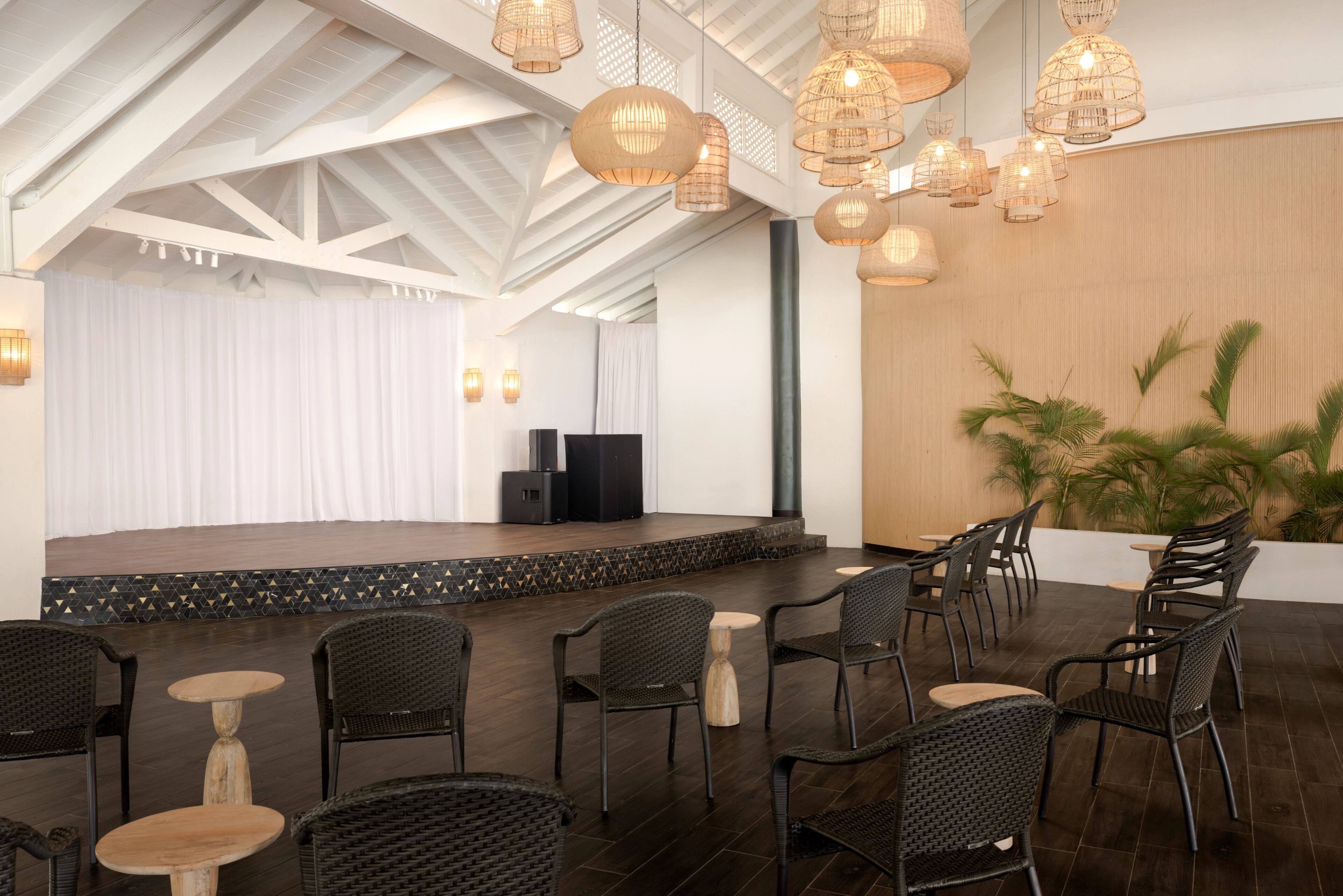 An indoor venue with a stage, black wicker chairs, and hanging wicker lamps.