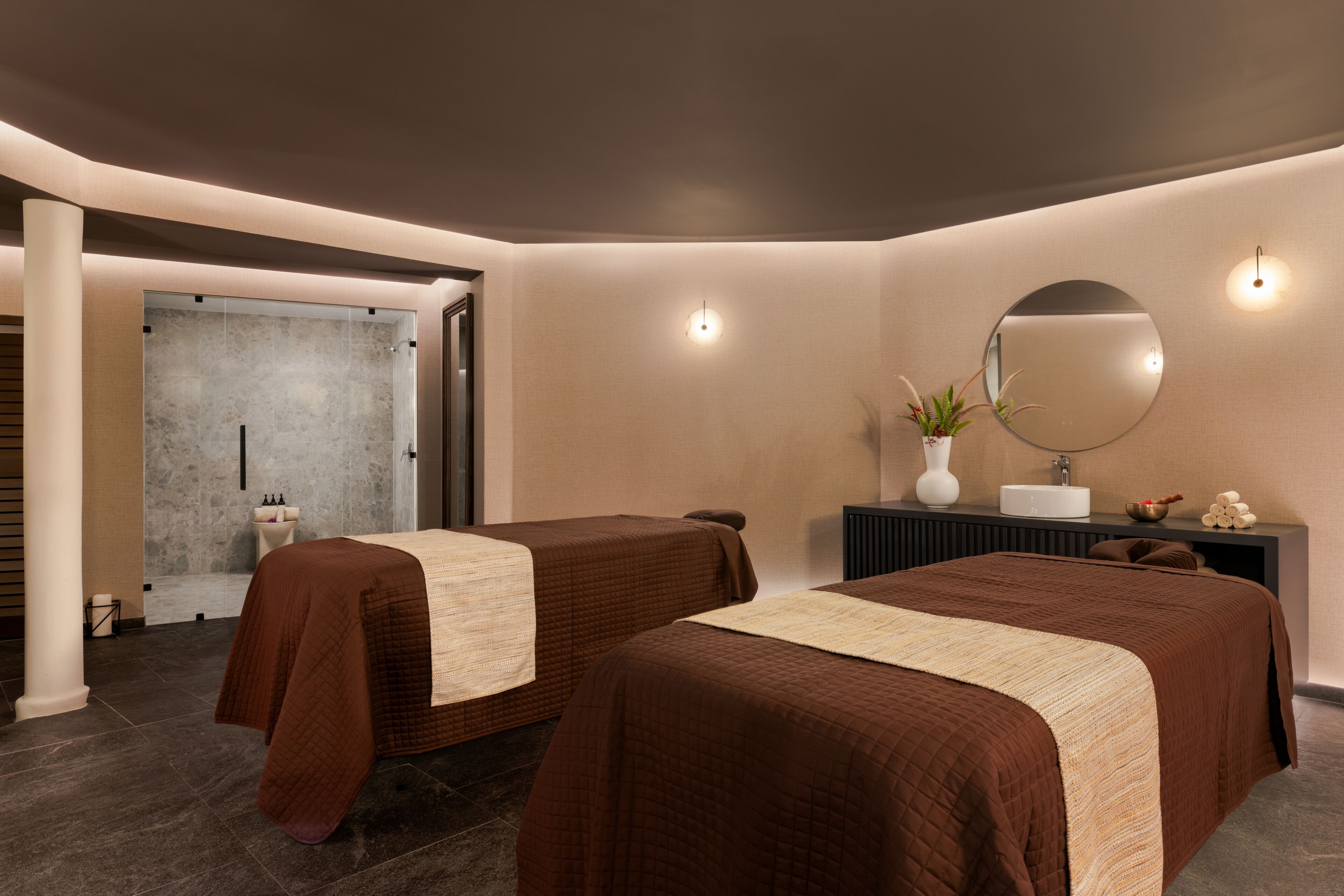 Une chambre d’hôtel moderne avec deux lits, un grand miroir, une coiffeuse avec un lavabo et une petite plante.
