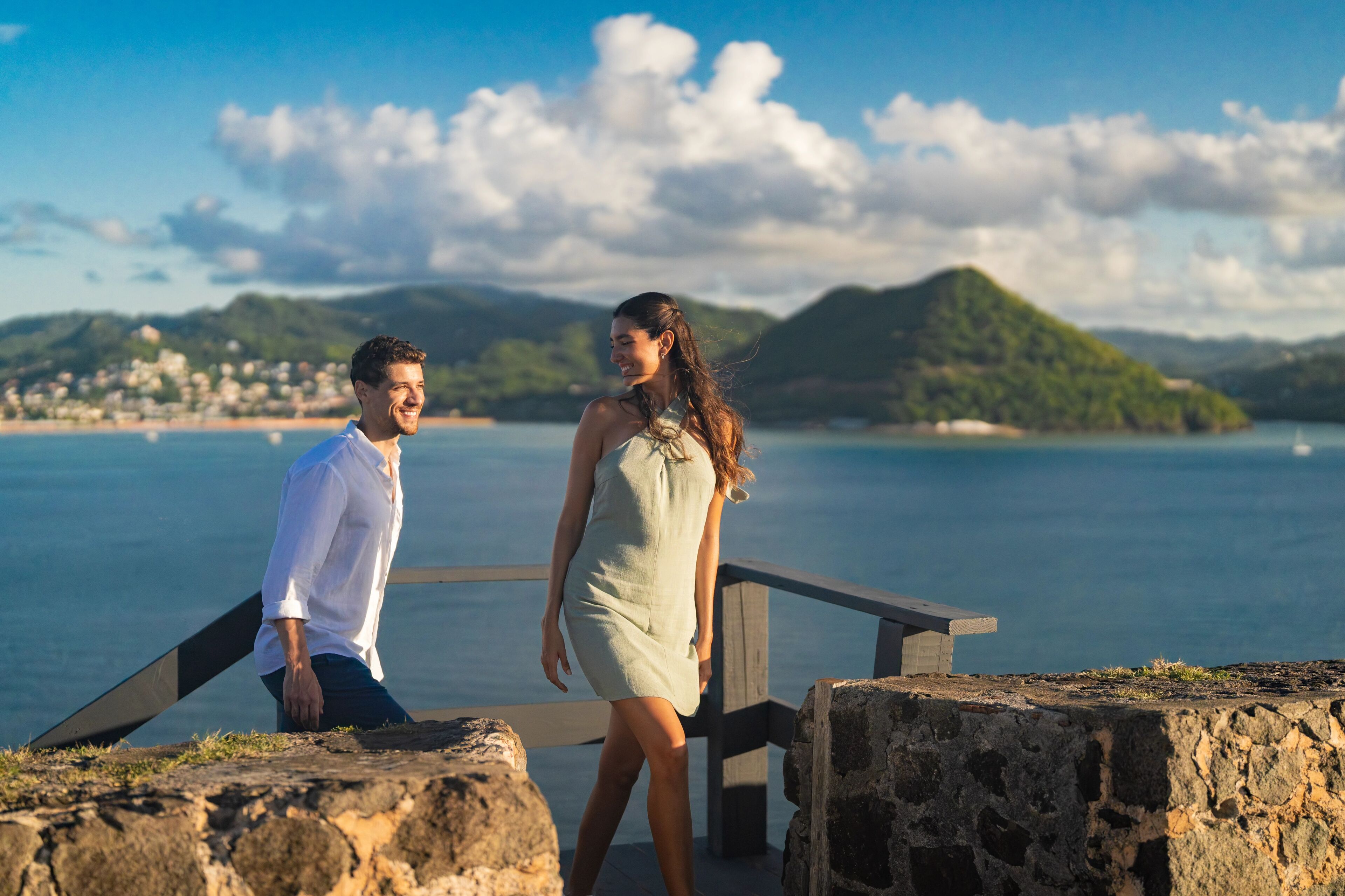 Un couple se tient sur une plateforme en pierre, protégée par une rambarde, et surplombe un paysage côtier composé de collines et d’une étendue d’eau.
