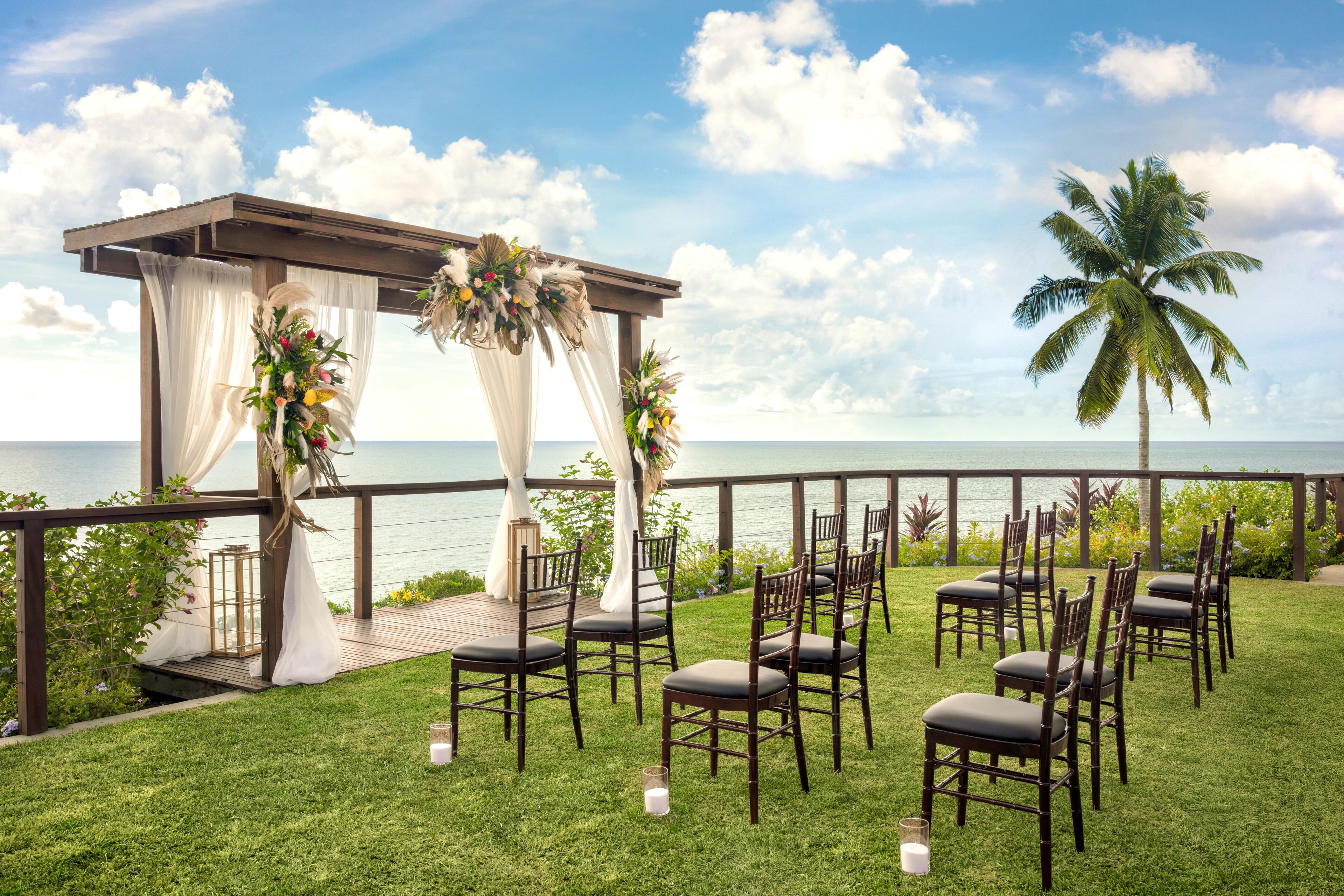 An outdoor wedding setup with a wooden pergola, white drapes, floral arrangements, and chairs arranged on a grassy area with a view of the ocean.