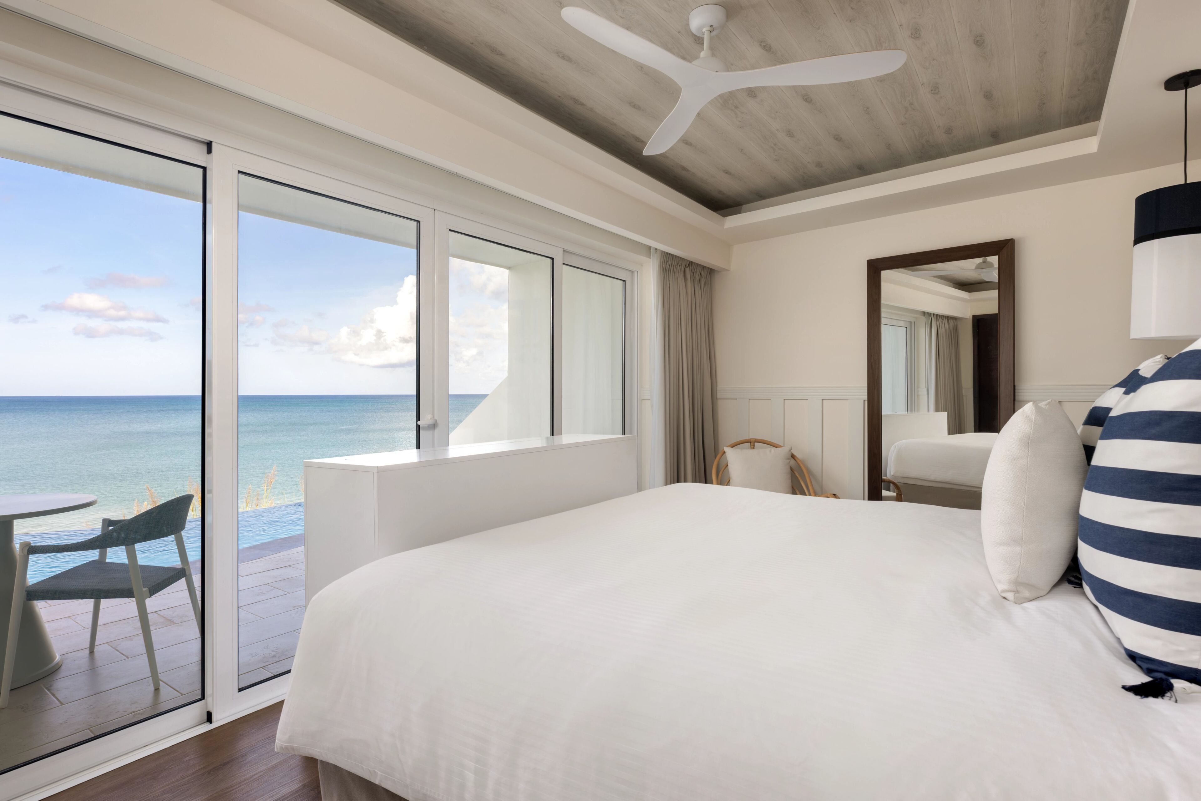 A hotel room with a large bed, a ceiling fan, a view of the ocean, and a balcony with a table and chair.