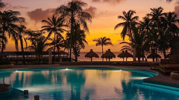 Ein Pool mit Sonnenuntergangsblick, umgeben von Palmen und Strohdächern.