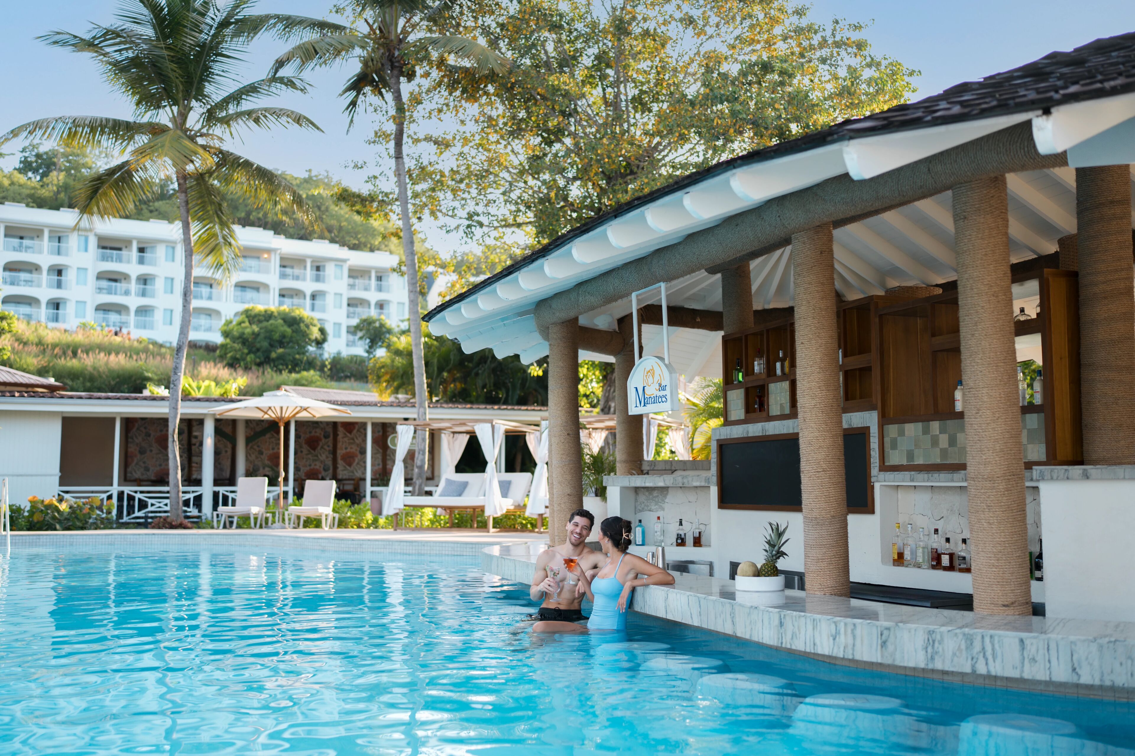 Una piscina con sillones de descanso, una zona de bar y un edificio con varios balcones al fondo.