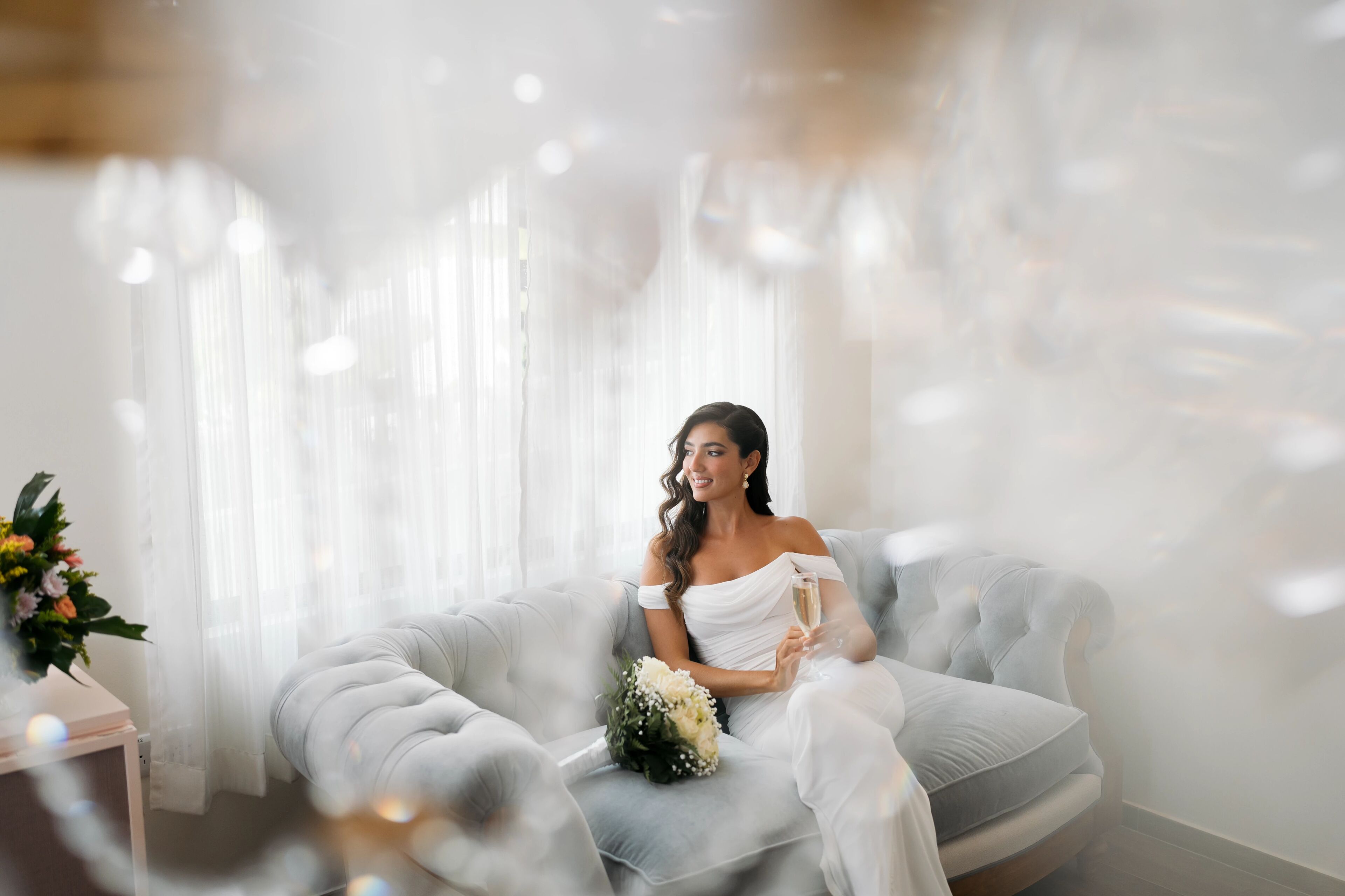 A person seated on a grey sofa in a room with sheer curtains and a floral arrangement.