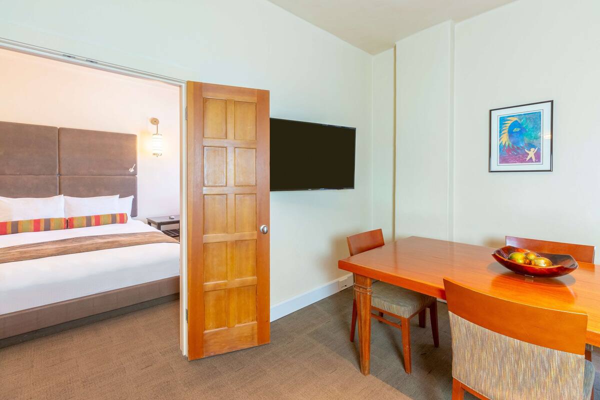 A hotel room with a bed, a dining table with chairs, a flat-screen TV mounted on the wall, and a framed picture.