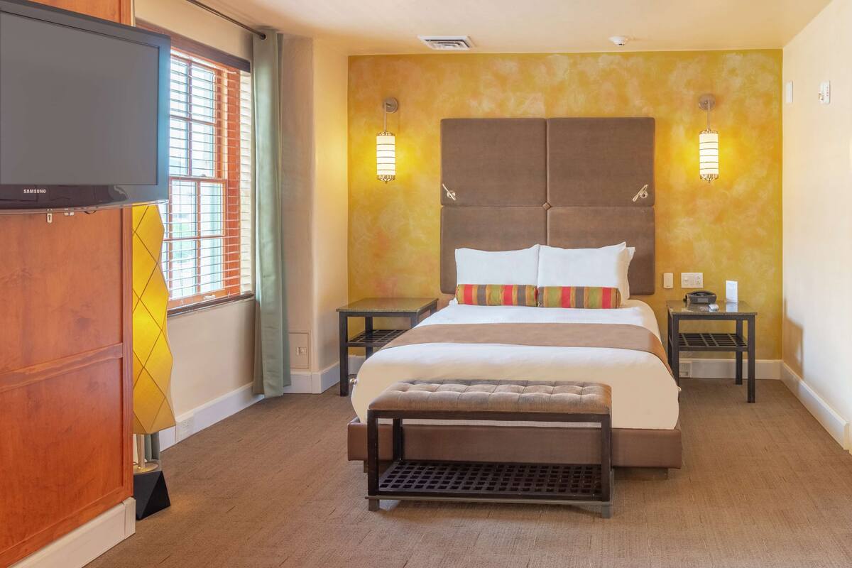 A hotel room with a large bed, two bedside tables, a bench, a TV, and a window with blinds.