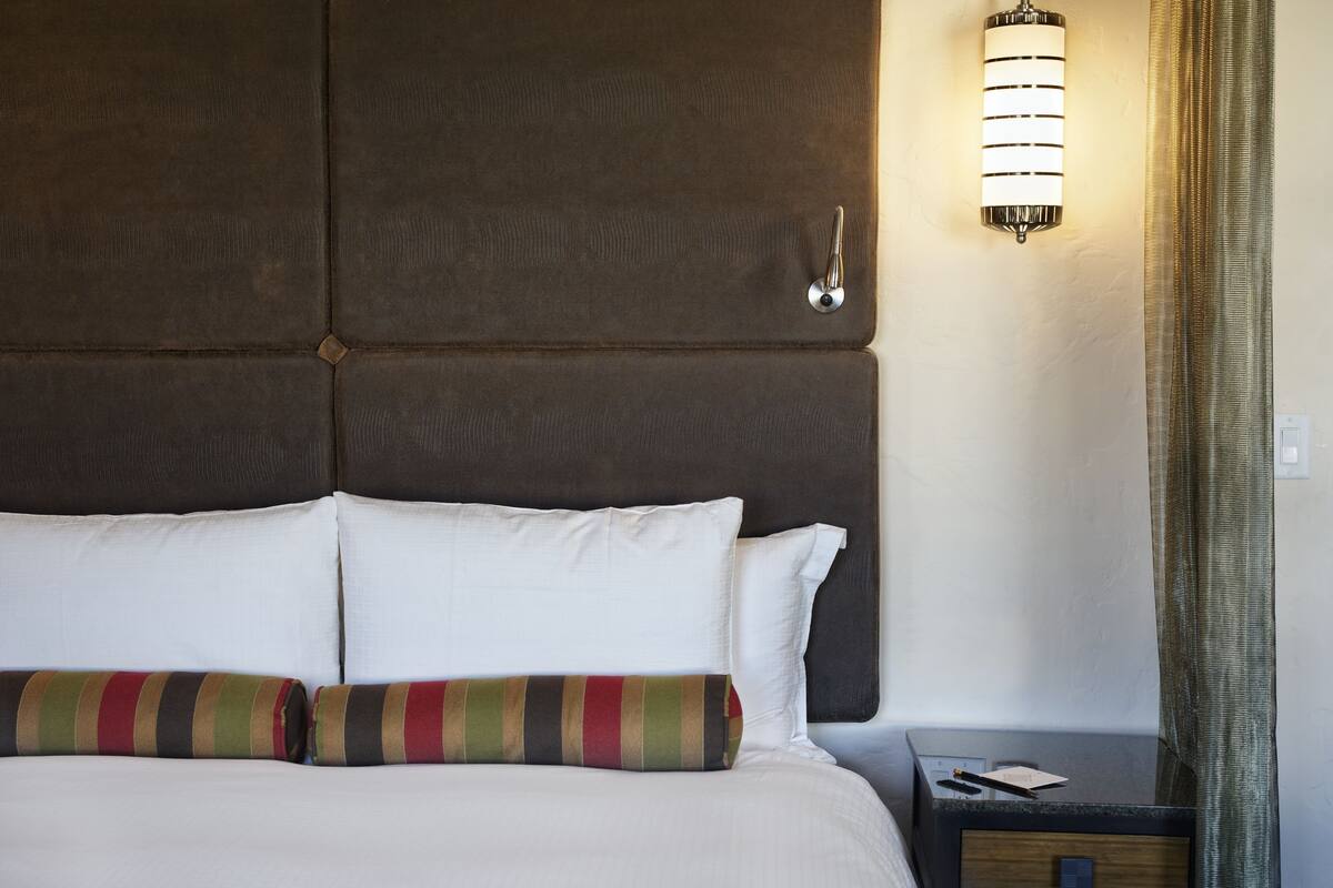 A hotel room with a large padded headboard, a bedside table with a lamp, and a striped throw pillow.