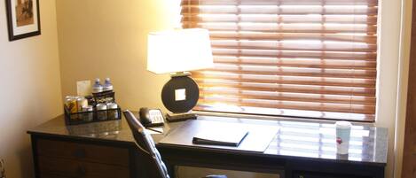 A hotel room with a desk, chair, lamp, and window with blinds.