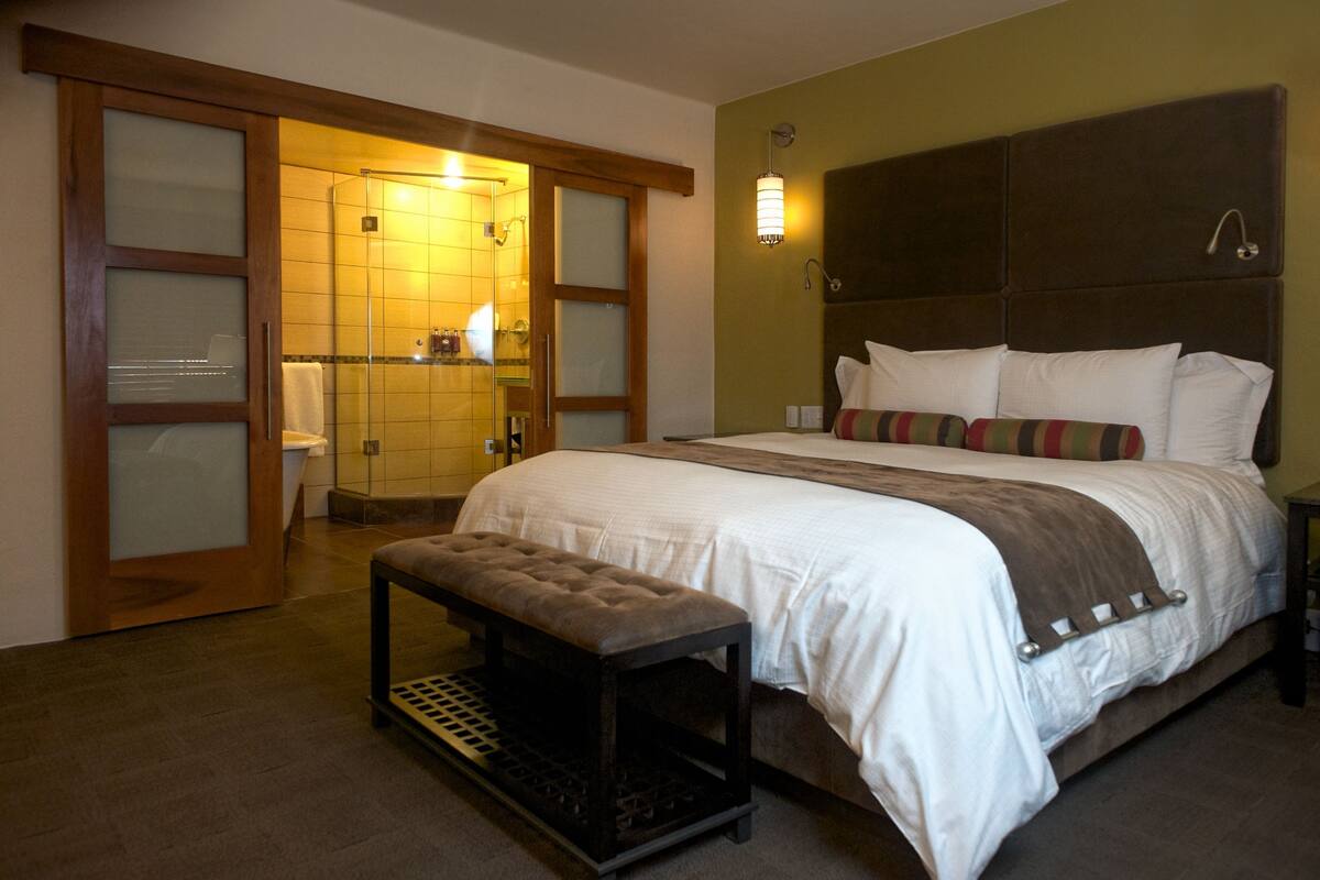 A bedroom with a large bed, a bench, a shower area, and a wall-mounted light fixture.