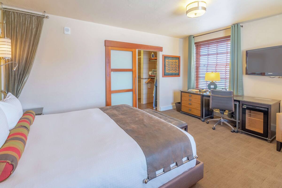 A hotel room with a large bed, a desk, a chair, a television, and a window with blinds.