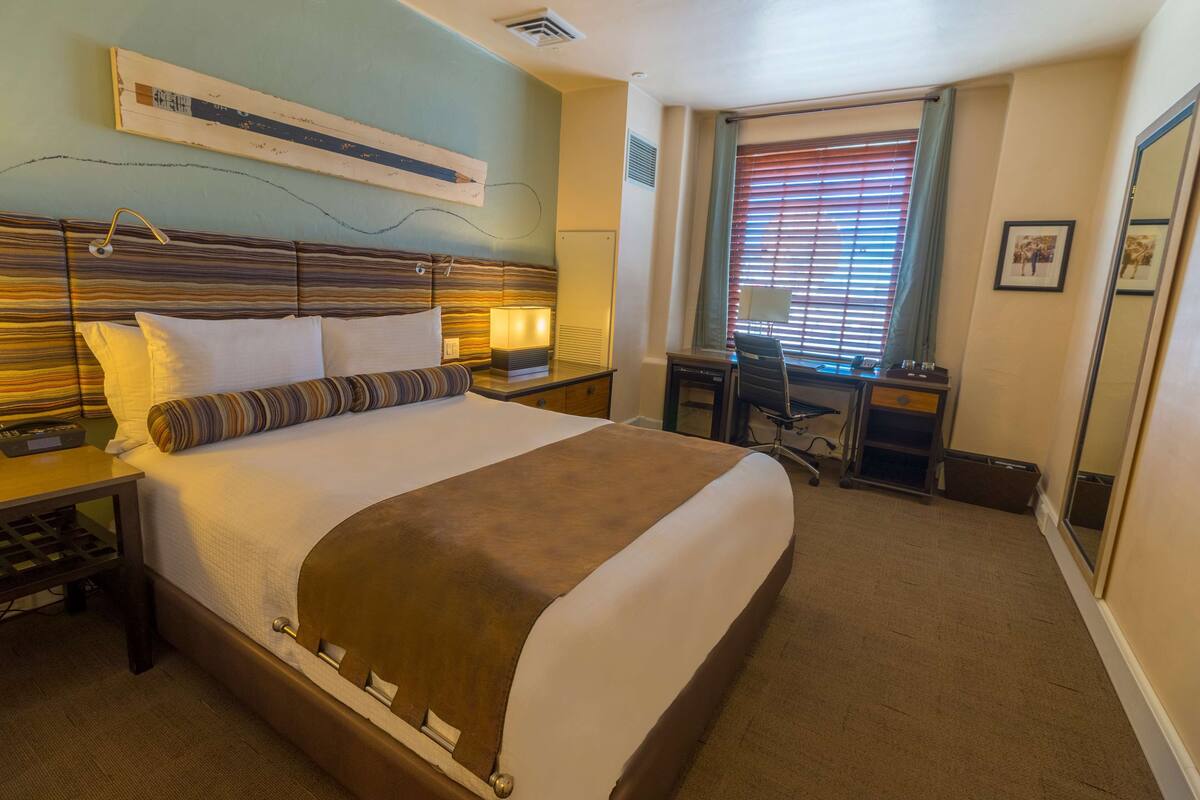 A hotel room with a large bed, bedside tables, a desk, and a window with blinds.