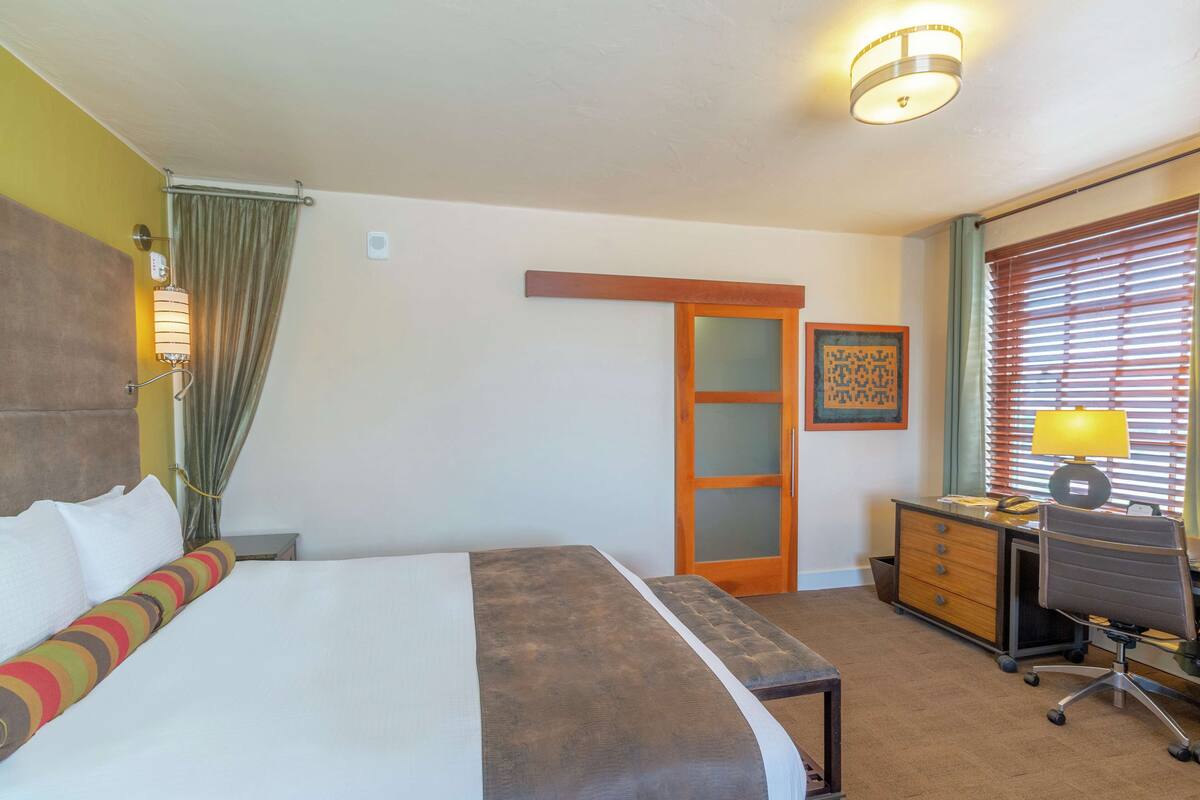 A hotel room with a large bed, a desk with a chair, a wardrobe, and a window with blinds.