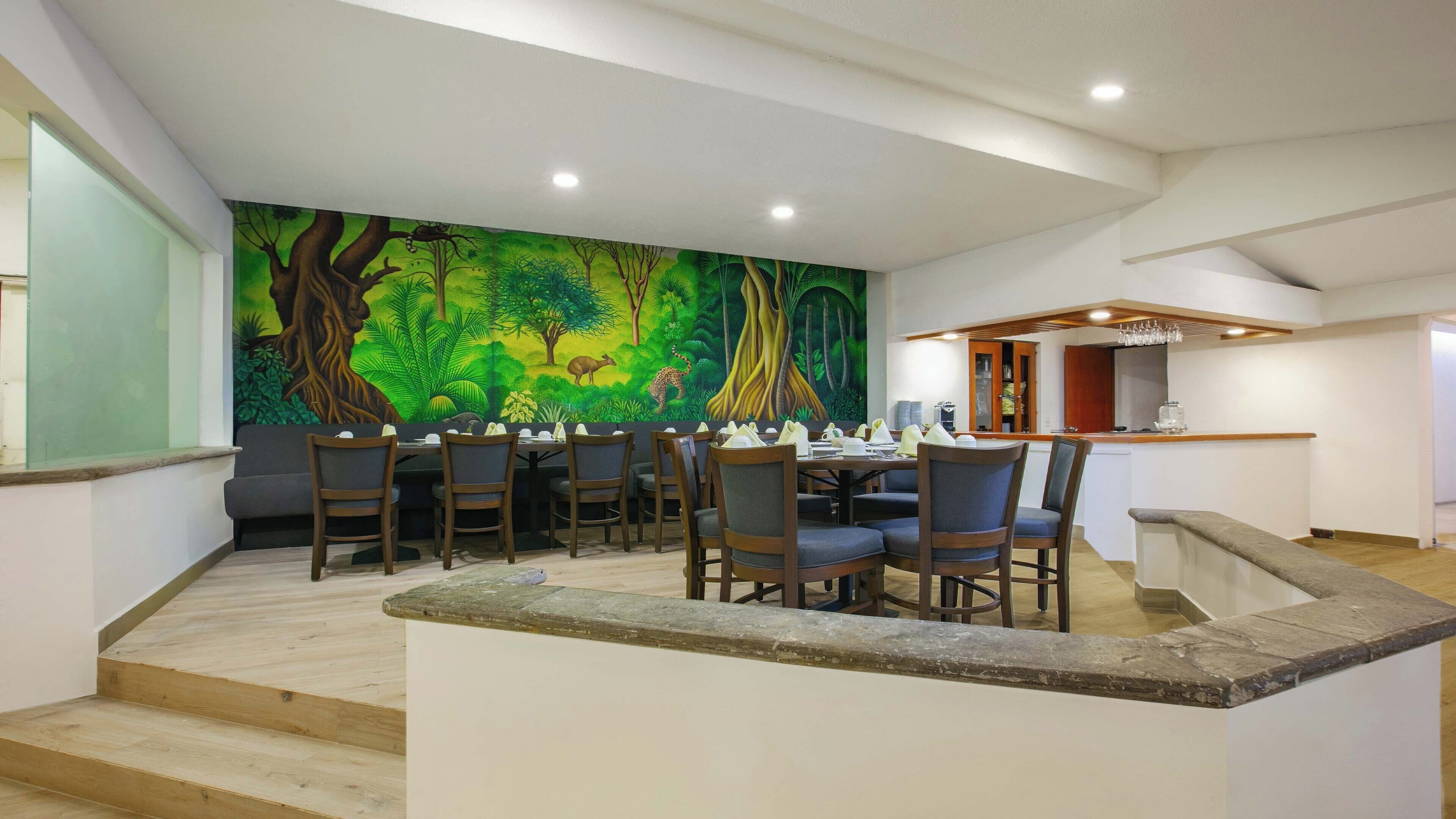A dining area with a mural of a forest scene, wooden chairs, and a small kitchen area with a counter and stools.