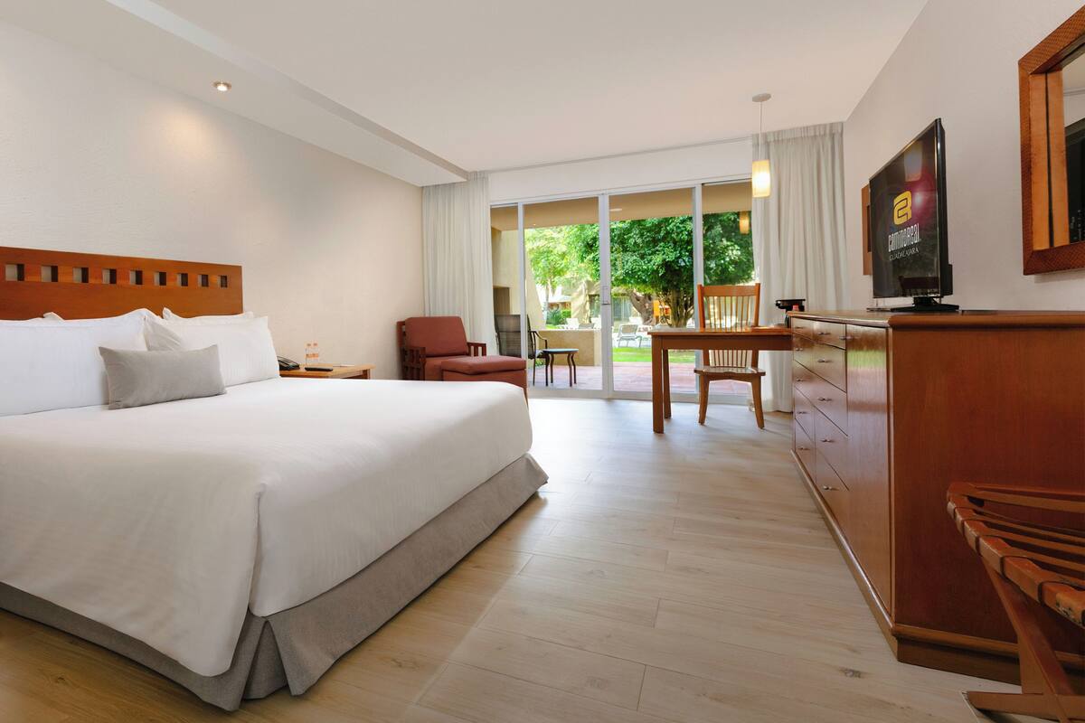 Ein Hotelzimmer mit einem großen Bett, einem Holzkopfstück, einem Schreibtisch mit Stuhl, einem Fernseher und Blick ins Freie.
