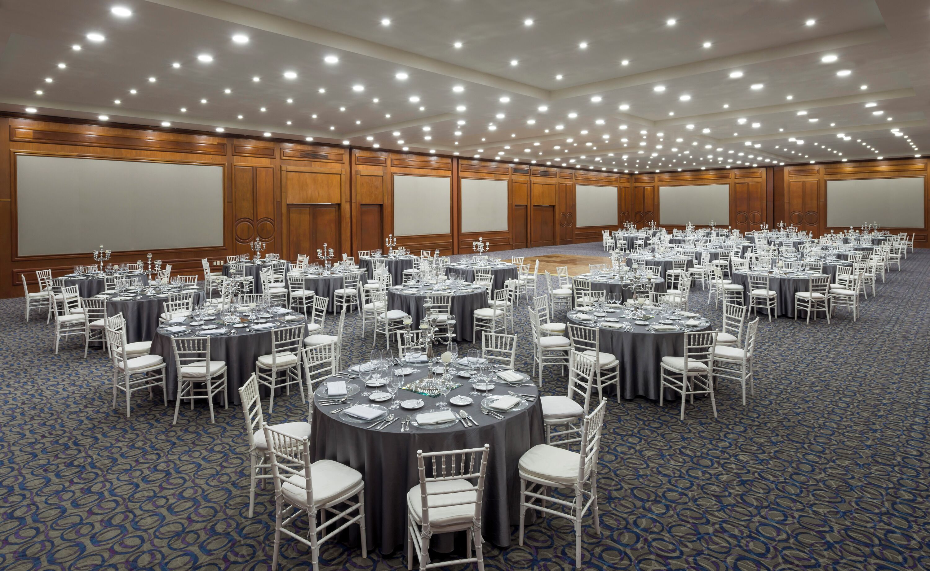 A large banquet hall with round tables set for dining, white chairs, and a carpeted floor.