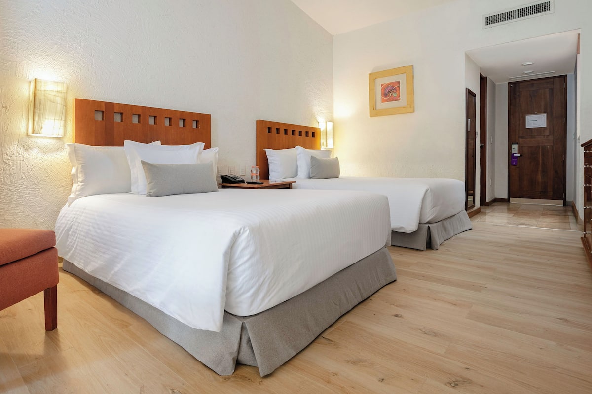 Ein Hotelzimmer mit zwei Betten, einem Holzkopfteil, einem Nachttisch mit Telefon und Wasserflasche, einer Holzbank und einem Holzschrank.