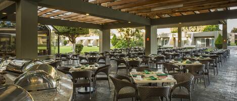 Un área de comedor al aire libre, techada, con varias mesas y sillas, una barra de buffet y vistas a un jardín ajardinado y edificios al fondo.