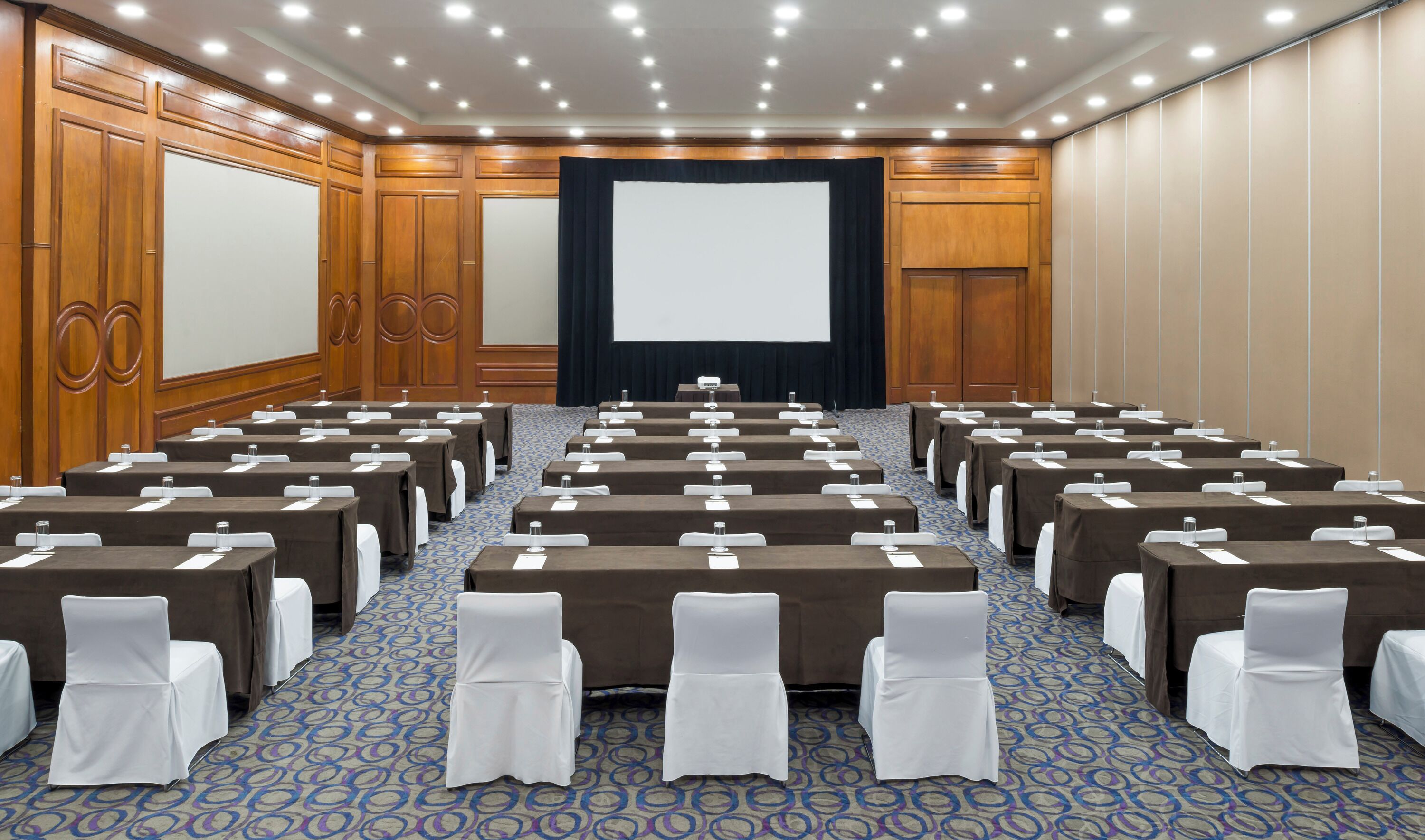 A conference room with rows of tables, white chairs, and a large screen at the front.